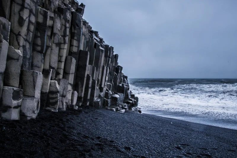 Hexagonal basalt columns on Reynisfjara Black Sand Beach in Iceland with waves crashing on the shore, showcasing dramatic volcanic formations.