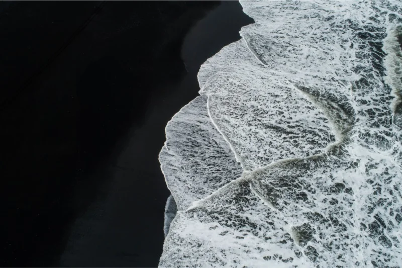 Aerial view of powerful waves crashing onto the volcanic black sand of Reynisfjara Beach in Iceland, showcasing nature's raw beauty and danger.