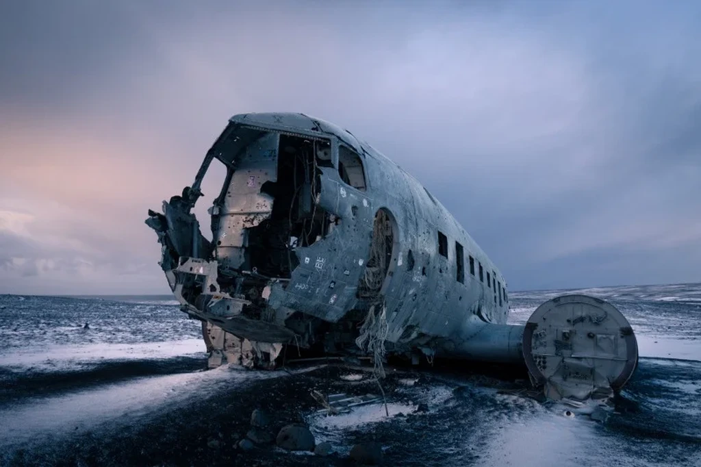 The wreckage of a DC-3 airplane lies on the snowy sands of Sólheimasandur Beach in Iceland. The front of the fuselage is partially disintegrated, revealing the hollow interior of the once-mighty aircraft. The weathered metal surface is marked by patches of rust and peeling paint, testifying to the harsh Icelandic elements it has endured. The surrounding landscape is desolate, with a pale sky and a hint of the ocean in the distance, creating a stark and hauntingly beautiful scene.