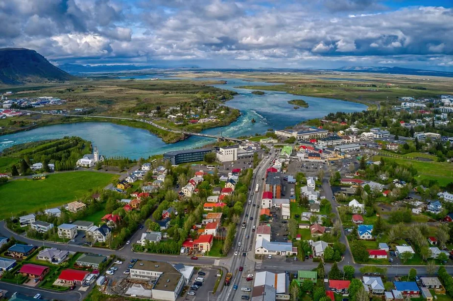 An aerial view of a small town in Iceland, showcasing a mix of colorful houses and buildings along a main road. The town is surrounded by lush green fields and winding blue rivers, with the landscape extending into the distance. The sky is partly cloudy, with patches of sunlight illuminating the vibrant scenery. The river winds through the town, adding to the picturesque setting. The image captures the peaceful and scenic environment typical of Icelandic towns, with a blend of natural beauty and human settlement