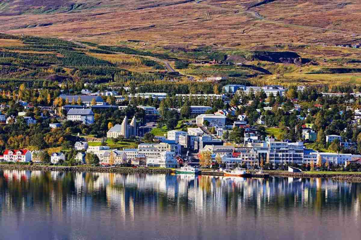 A picturesque view of Akureyri, the largest town in northern Iceland, nestled along the coastline of Eyjafjörður. The town's charming buildings, including traditional Icelandic houses and modern structures, are vividly reflected in the calm fjord waters. In the foreground, colorful rooftops stand out amidst a backdrop of lush greenery and autumnal hues, while further behind, the landscape rises into rolling hills. The scene captures the serene beauty of Akureyri, highlighting its unique blend of nature and urban life