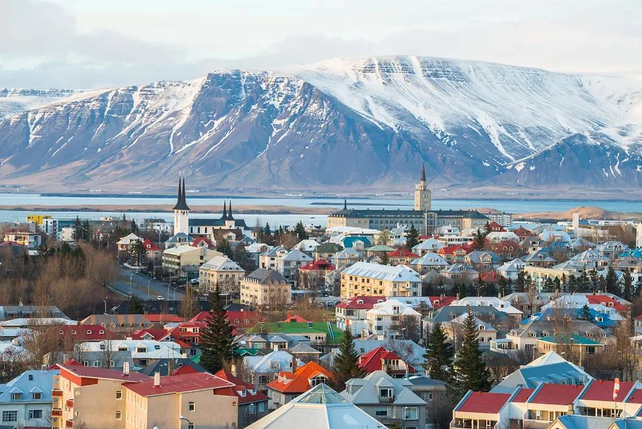 A vibrant cityscape of Reykjavik, Iceland, featuring colorful houses with red, green, and blue roofs, set against a backdrop of snow-capped mountains. The city is dotted with spires, including the iconic Hallgrímskirkja church. The mountains, partially covered with snow, create a stunning contrast with the bright buildings below. The calm waters of the nearby bay reflect the serene winter landscape. The overall scene captures the unique blend of urban life and natural beauty in Iceland's capital