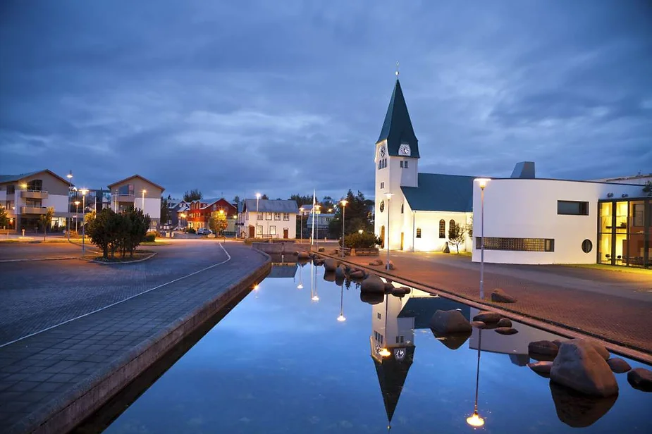 A serene evening view of Akureyrarkirkja, the church in Akureyri, Iceland, beautifully illuminated against a dusky sky. The church's distinctive green spire and white facade stand out, reflected in a calm pond nearby. Surrounding buildings are softly lit, adding a warm glow to the quiet scene. The tranquil water mirror doubles the charm of this peaceful townscape, capturing the calm atmosphere of Akureyri as the day transitions to night