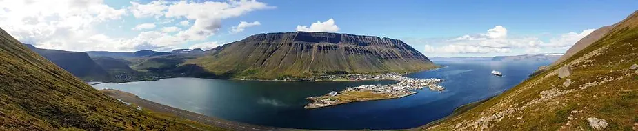 A panoramic view of a serene Icelandic fjord, surrounded by rugged mountains. A small town sits nestled along the coastline, with a few scattered buildings and a harbor. The fjord's deep blue waters contrast with the green and brown tones of the surrounding hillsides. The clear sky above is dotted with a few fluffy clouds, adding to the tranquil beauty of the scene. This image captures the stunning natural landscape and peaceful atmosphere of Iceland's coastal regions