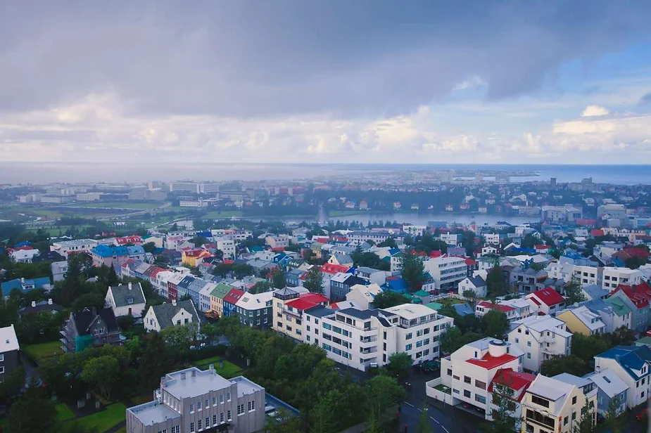 A panoramic view of Reykjavik, Iceland's capital, on a cloudy day. The cityscape features colorful rooftops in shades of red, blue, yellow, and white, set against a backdrop of lush greenery and urban buildings. The Atlantic Ocean is visible in the distance, with a light mist creating a serene atmosphere. The image captures the charm of Reykjavik's architecture and the city's unique blend of natural and urban landscapes