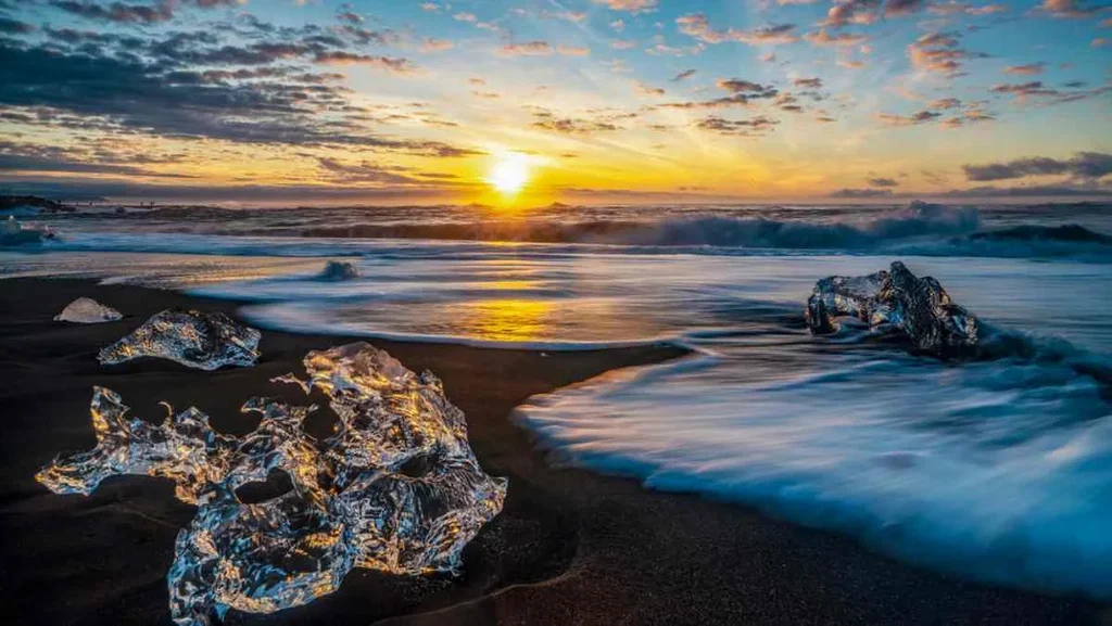 A stunning view of Diamond Beach in Iceland during sunset. The beach is famous for its striking black sand and the glistening ice chunks scattered along the shore, which resemble diamonds. The sun is setting on the horizon, casting a golden glow over the ocean waves and illuminating the ice formations. The sky is a mix of warm orange and pink hues, reflecting beautifully off the water and ice. The smooth waves gently wash over the beach, creating a serene and mesmerizing landscape. Diamond Beach is renowned for its unique beauty and the contrasting elements of ice, sand, and water.