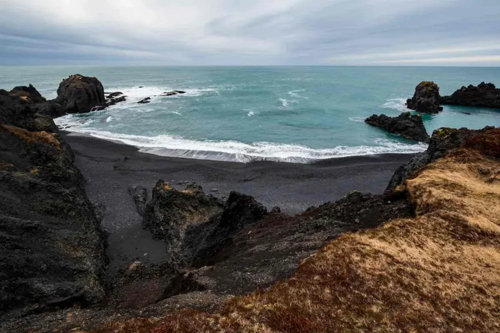 A rugged coastline view of Djupalonssandur Beach in Iceland, characterized by its distinctive black sand. The ocean waves gently lap against the shore, framed by jagged rock formations that jut into the water. The sky is overcast, casting a moody and dramatic atmosphere over the landscape. The black sand contrasts sharply with the surrounding rocky cliffs and the distant, churning sea. This beach, known for its unique geological features and historical significance, offers a glimpse into the natural beauty and wild landscapes that Iceland is renowned for.