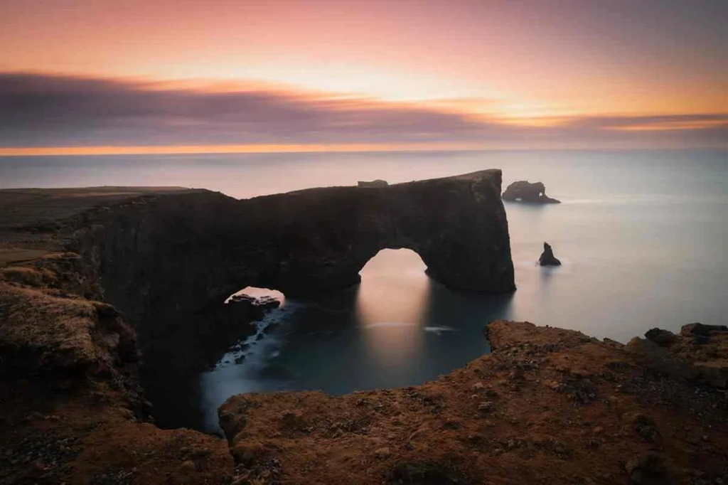 A breathtaking sunset illuminates the iconic Dyrhólaey arch, a natural rock formation along the southern coast of Iceland. The arch stands tall and majestic over the calm waters of the Atlantic Ocean. The sky is painted in shades of orange, pink, and purple as the sun sets, casting a warm glow over the scene. The rugged cliffs and unique shape of the arch create a striking silhouette against the vibrant sky. This serene and picturesque landscape captures the stunning beauty of Iceland's coastline and the tranquility of the moment.