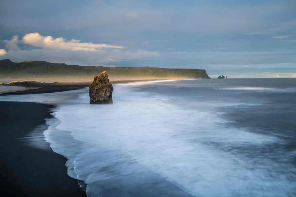 A panoramic view of Víkurfjara Black Sand Beach in Iceland, with waves crashing onto the shore under a cloudy sky. The beach's distinctive black sand contrasts sharply with the surrounding landscape, including the grassy cliffs in the foreground and the distant cliffs along the coast. The famous Reynisdrangar rock formations can be seen jutting out of the ocean in the distance. The scene captures the dramatic and rugged beauty of Iceland's coastline, characterized by its volcanic origins and dynamic weather patterns. The ocean appears powerful and untamed, embodying the wild essence of this iconic Icelandic location.