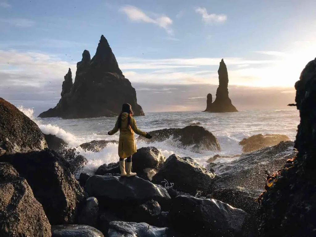 A person stands on the rocky shore of Reynisfjara Beach in Iceland, facing the ocean and the dramatic sea stacks known as Reynisdrangar. The individual, dressed in a yellow coat and hat, gazes out at the towering, jagged rock formations rising from the turbulent sea. The waves crash against the dark basalt rocks at the person's feet, sending sprays of water into the air. The sky above is partly cloudy with a hint of sunlight breaking through, casting a warm glow over the scene. The image captures the raw power and unique beauty of this iconic Icelandic coastal landscape.