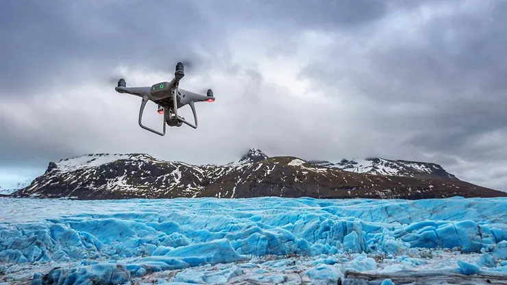 A drone flying over a bright blue glacier with snow-covered mountains in the background in Iceland.