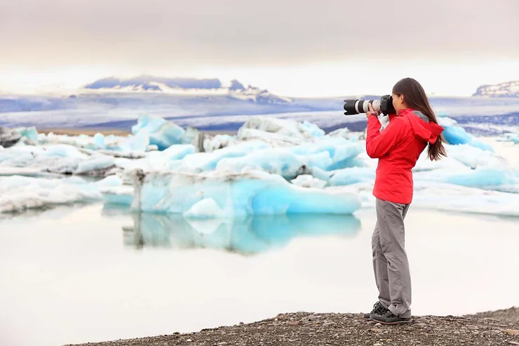 A woman in a red jacket photographing icebergs in a glacial lagoon in Iceland with a professional camera.