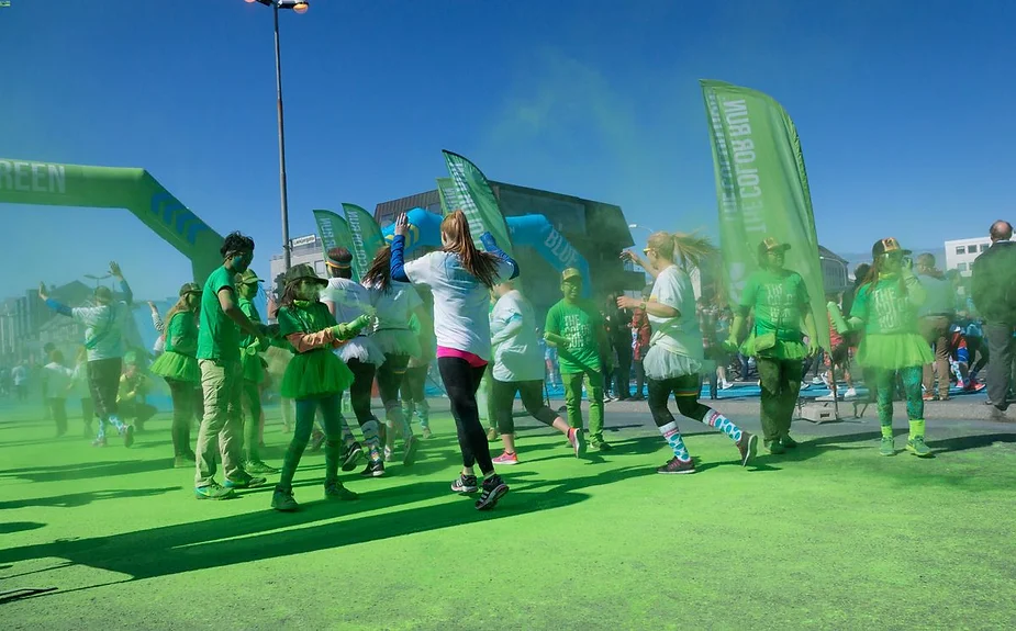 Participants at a color run event are captured mid-celebration, covered in green powder. The image shows a lively scene with runners and walkers dressed in green outfits and tutus, some in white shirts, moving through a cloud of green powder. The event takes place under a clear blue sky, with green flags and an archway visible in the background. The ground is also covered in green powder, contributing to the vibrant, festive atmosphere. The participants appear joyful and energetic, enjoying the colorful and fun nature of the event