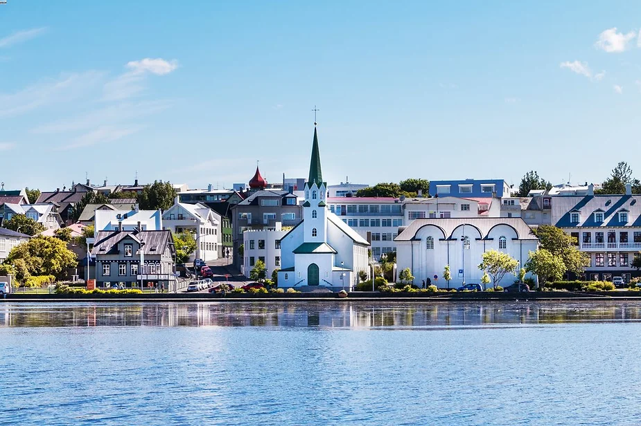 A picturesque view of Reykjavik's cityscape features the iconic Fríkirkjan church with its green spire, located by the serene Tjörnin lake. The image captures a bright, sunny day with clear blue skies. The buildings along the waterfront display a variety of architectural styles, from traditional Icelandic houses to modern structures. The calm water of Tjörnin reflects the buildings and sky, adding to the tranquil atmosphere of the scene. The lush greenery and well-maintained surroundings contribute to the city's charming and inviting appearance