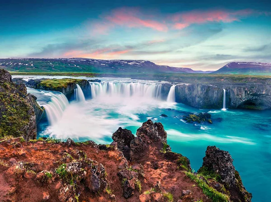 A stunning view of Goðafoss Waterfall in Iceland, known for its semi-circular shape and powerful, cascading waters. The waterfall plunges into a turquoise blue pool surrounded by rugged, rocky cliffs. The foreground features a reddish-brown, rocky outcrop with patches of green vegetation. In the background, expansive, snow-capped hills stretch under a dramatic sky tinged with shades of pink and blue, suggesting either dawn or dusk. The scene captures the natural beauty and raw power of one of Iceland's most iconic waterfalls