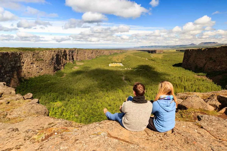 A couple sits on the edge of Asbyrgi Canyon in Iceland, enjoying a panoramic view of the lush, green valley below. The canyon walls rise steeply on either side, creating a horseshoe-shaped formation. The couple, dressed warmly, gazes out over the vast expanse of the canyon, with a bright, partly cloudy sky above. The landscape is covered in dense green vegetation, extending into the distance under a vibrant blue sky