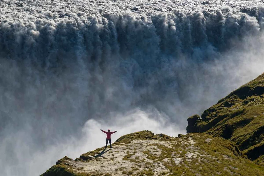 A person stands near the edge of a cliff at Dettifoss, one of Europe's most powerful waterfalls, located in Iceland. The individual, wearing a bright red jacket, raises their arms in awe and exhilaration. Behind them, an immense and roaring wall of water cascades over the cliff, creating a misty spray and highlighting the sheer force of the waterfall. The surrounding landscape is rugged and green, contrasting with the dramatic water flow