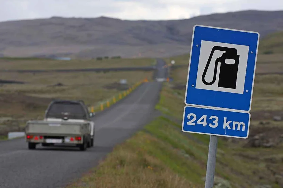 A road sign indicating a gas station 243 kilometers away on a rural stretch of Iceland's Ring Road. The sign, featuring a gas pump icon, is blue with white text. In the background, a vehicle travels along the winding road, which stretches into the distance with mountains and open countryside on either side. The landscape is predominantly brown and green, under a cloudy sky