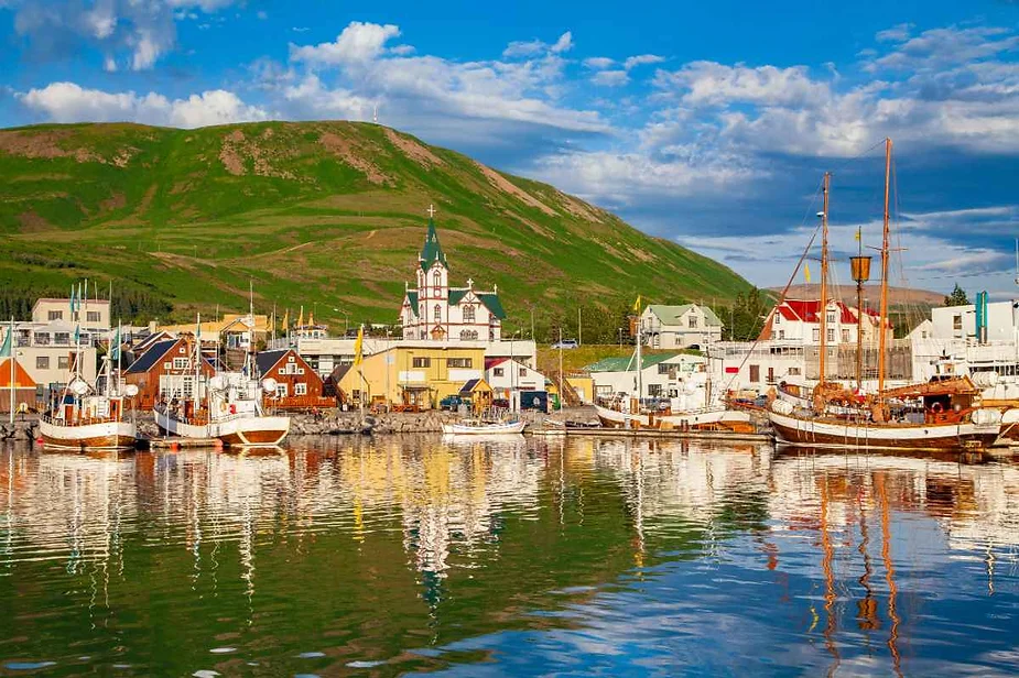 A picturesque scene of Húsavík Harbor in Iceland, featuring a collection of colorful buildings along the waterfront. Traditional Icelandic fishing boats are docked along the harbor, reflecting on the calm, clear water. A charming church with a distinctive spire stands prominently among the buildings, adding to the quaint, coastal village atmosphere. In the background, lush green hills rise gently, under a bright blue sky with scattered clouds, creating a beautiful and serene landscape