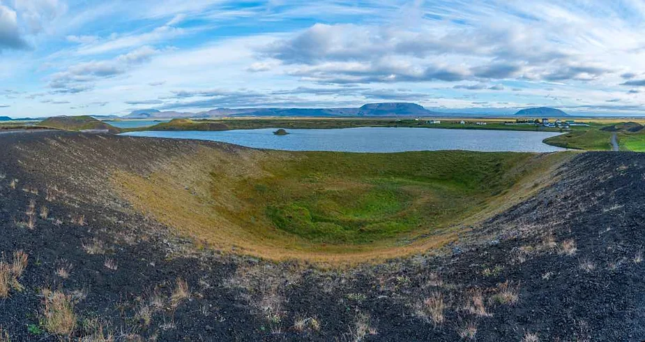 A panoramic view of a volcanic crater and lake in Iceland. The crater, with its dark rocky rim, surrounds a lush, green floor. In the distance, a calm blue lake stretches out, bordered by gentle hills and distant mountains. The sky is partly cloudy, with patches of blue visible, and the landscape features a mix of green grass and volcanic rock. The peaceful, expansive scenery highlights the unique geological features of Iceland