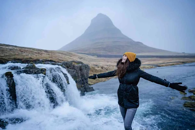 Woman standing with arms outstretched near a waterfall, with Kirkjufell mountain in the background, enjoying the winter scenery in Iceland.