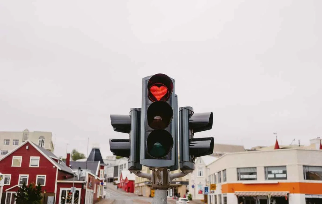 A unique traffic light in Akureyri, Iceland, displaying a red heart-shaped stop signal. The background shows a quaint street lined with colorful buildings, including a red house with white trim and other pastel-colored structures. The sky is overcast, giving a soft, diffused light to the scene. This heart-shaped traffic signal is a distinctive feature in Akureyri, symbolizing the town's warmth and friendly atmosphere.