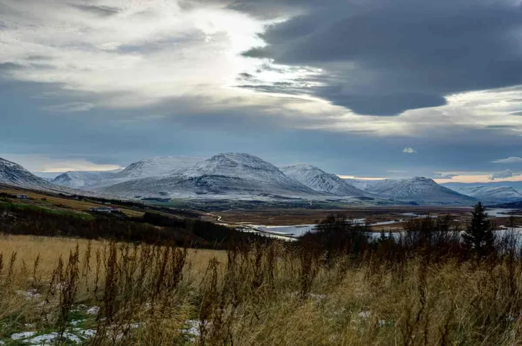 A scenic view of Mount Sulur in Akureyri, Iceland, with snow-capped peaks and lush hills in the foreground.