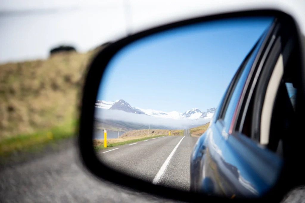 A scenic view from a car's side mirror, showing a winding road in Akureyri leading to snow-capped mountains.