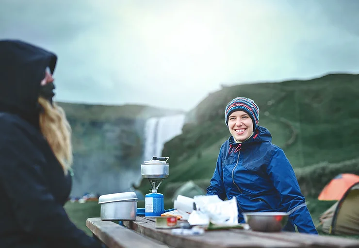 Two people enjoying a camping trip near Skógafoss waterfall in Iceland, with camping gear and a portable stove on a wooden table, surrounded by green hills and mist.