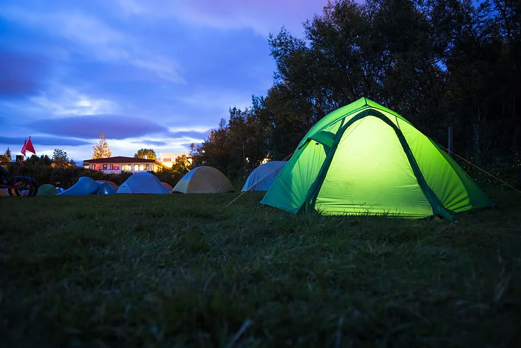 Illuminated green tent at a campsite in Iceland during twilight, with other tents and a lit building in the background.