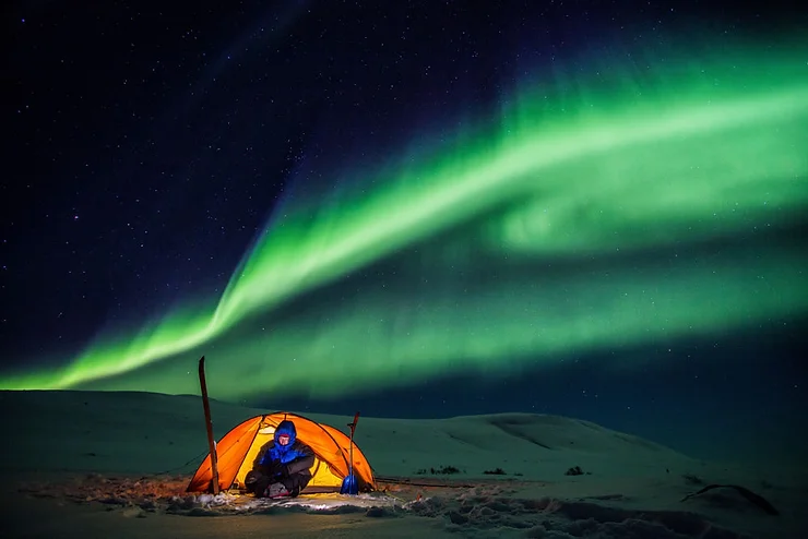 Person sitting in a tent on a snowy landscape with the vibrant Northern Lights illuminating the night sky in Iceland.