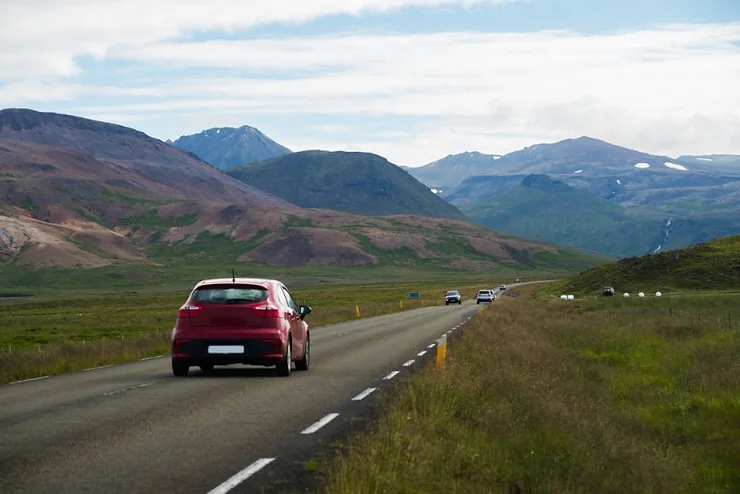 A red car driving along a scenic road in Iceland with mountains and green fields in the background.