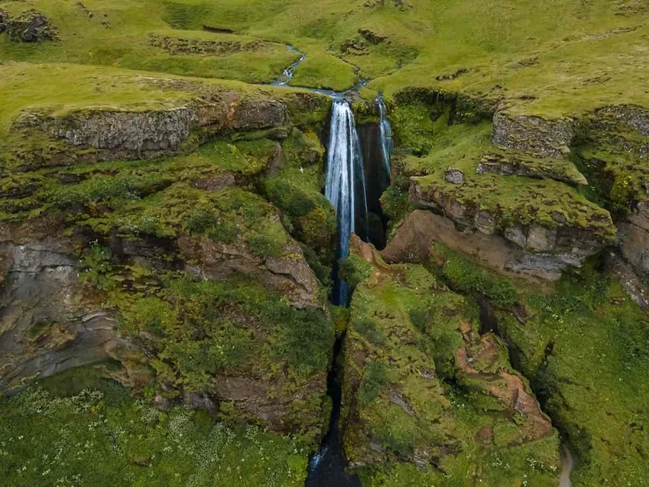 A breathtaking aerial view of a secluded waterfall cascading down a deep gorge surrounded by lush green landscape. The waterfall plunges from a narrow cliff into a narrow chasm, with moss-covered rock walls and verdant grasslands extending around it. The scene captures the natural beauty and serenity of this hidden gem, with the vibrant greenery contrasting against the darker rock formations. The water appears crystal clear as it descends, adding to the picturesque quality of the landscape