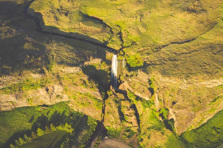 An aerial view of a picturesque landscape featuring a waterfall cascading down a cliff into a small basin. The waterfall is surrounded by a vibrant, lush green landscape, with the terrain showing various shades of green vegetation. The rocky cliffs are interspersed with patches of greenery, creating a natural, rugged beauty. The winding stream feeding the waterfall can be seen flowing through the terrain, with shadows and light playing on the landscape, highlighting its diverse textures and features. The image captures the serene and untouched beauty of nature from above