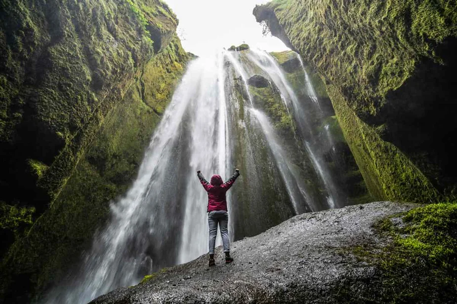 A person stands on a moss-covered rock platform, arms raised in awe, before a majestic waterfall cascading down a steep cliff. The waterfall's water flows powerfully from above, surrounded by lush green moss and vegetation clinging to the rocks. The scene captures a sense of wonder and the grandeur of nature, with the person dwarfed by the immense height of the waterfall. The water creates a misty atmosphere, adding to the ethereal quality of the landscape