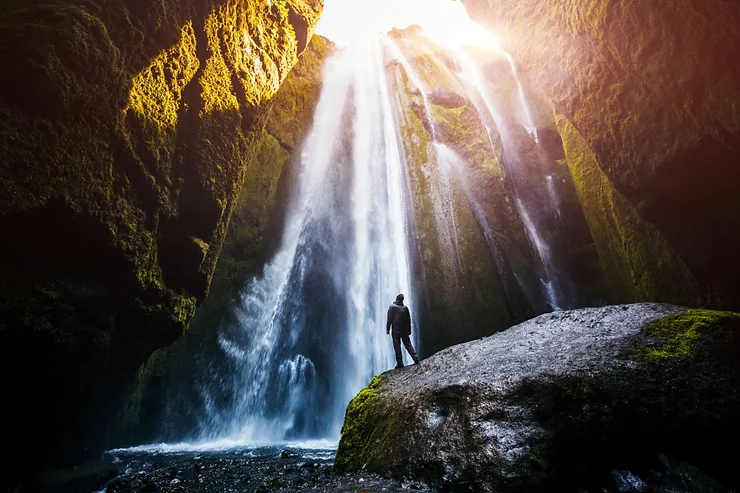 A person standing on a rock admiring the majestic Gljúfrabúi Waterfall, with sunlight streaming through the cascading water in Iceland.