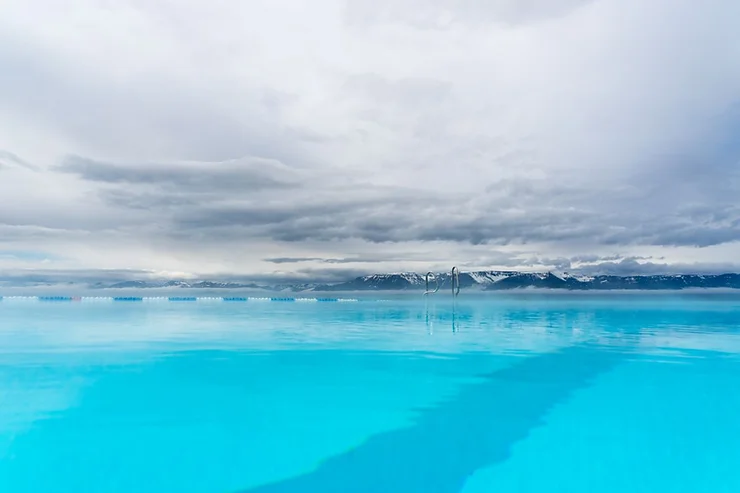 A serene view of the Hofsós Swimming Pool in Iceland, with crystal clear blue water reflecting the overcast sky and distant snow-capped mountains.