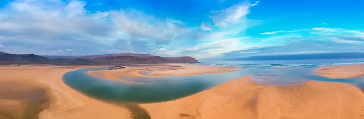 A panoramic view of Raudasandur Beach in Iceland, showcasing its unique red sand, winding river channels, and clear blue sky.