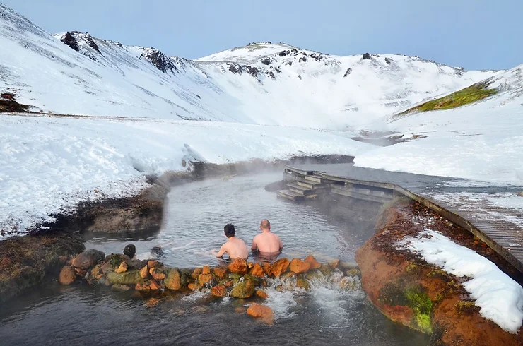 Two people enjoying a hot bath in the Reykjadalur Thermal River, surrounded by snowy mountains and steaming water in Iceland.