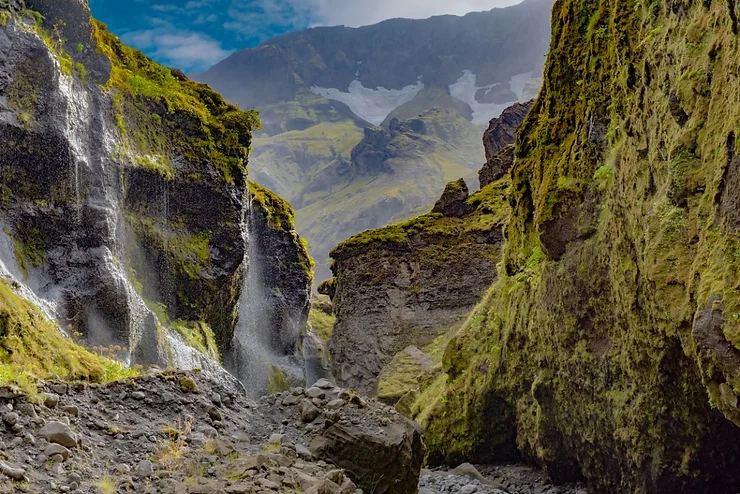 View of Stakkholtsgja Canyon in Iceland, featuring moss-covered cliffs and a cascading waterfall with majestic mountains in the background.