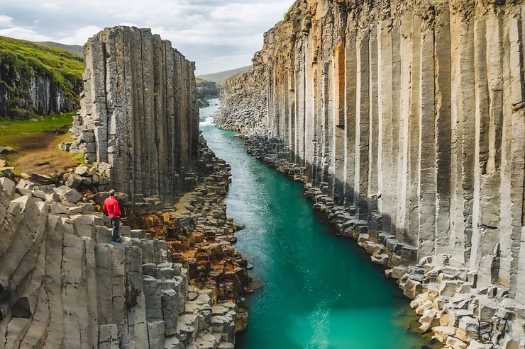 A person in a red jacket standing on the edge of the basalt column cliffs of Stuðlagil Canyon, with the turquoise river flowing through the canyon in Iceland.