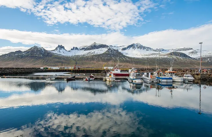 A serene harbor with boats docked and snow-capped mountains in the background, representing the weather and temperature in East Iceland.