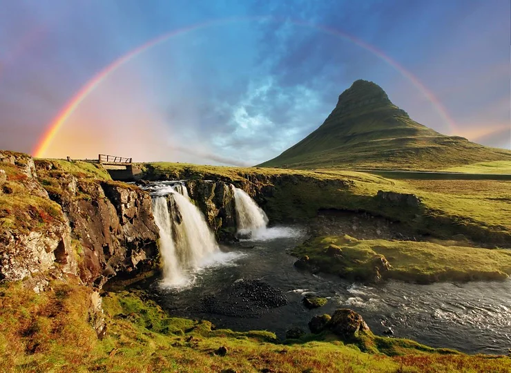 A vibrant landscape in Iceland featuring a waterfall, lush green hills, and a rainbow arching over Kirkjufell mountain, representing typical weather in July.