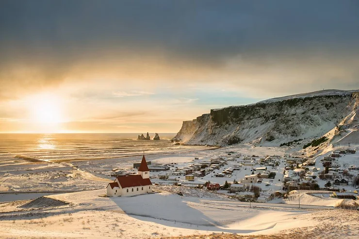 A snowy coastal village in Iceland with the sun setting over the ocean, typical of March weather.