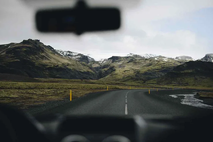 A scenic view through a car windshield of a winding road in Iceland, surrounded by rugged mountains partially covered in snow, under an overcast October sky.