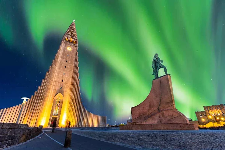 Hallgrimskirkja Church in Reykjavik, Iceland, lit up against the backdrop of vibrant green Northern Lights in October, with a statue in the foreground.