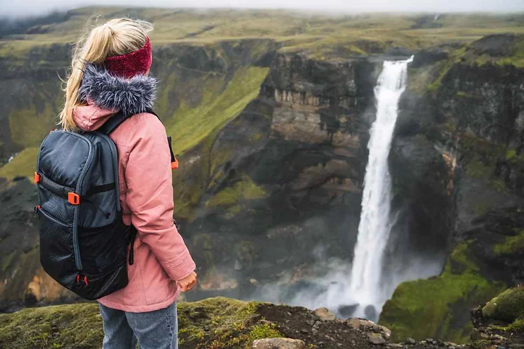 A woman with a backpack and winter headband standing on a cliff, overlooking a stunning waterfall surrounded by lush green cliffs in Iceland during October.