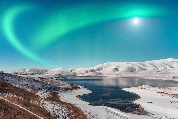 A mesmerizing view of the Northern Lights illuminating the October sky above a serene lake surrounded by snow-covered hills in Iceland.