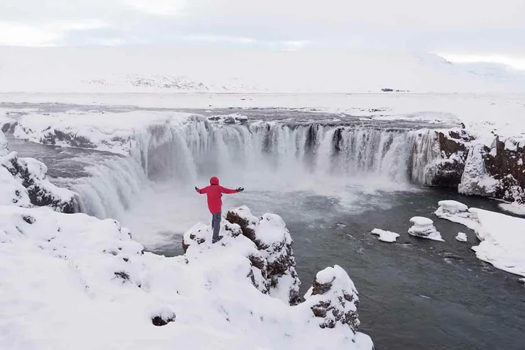 A person in a red jacket standing on a snow-covered cliff, arms outstretched, facing a majestic frozen waterfall surrounded by a snowy landscape in Iceland.