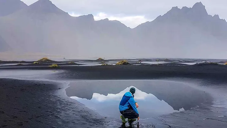 A person in a blue jacket and beanie crouching by a reflective pool on a black sand beach in Iceland, with misty mountains looming in the background under an overcast sky.